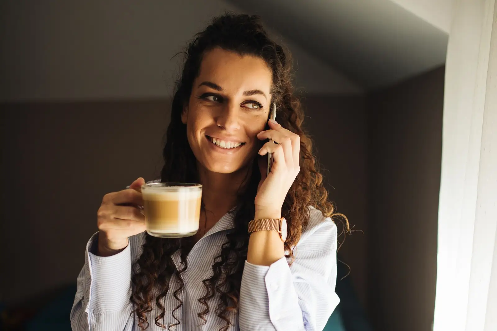 A young woman talking on the phone by a window, holding a coffee